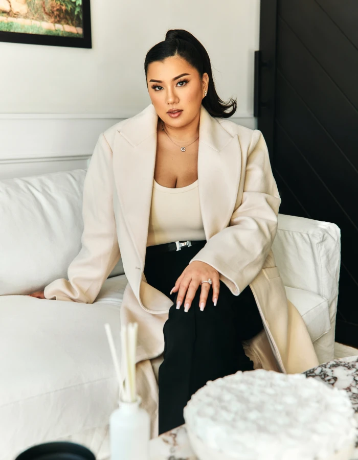 Remi slaying in a business long blazer jacket, sitting on a white couch